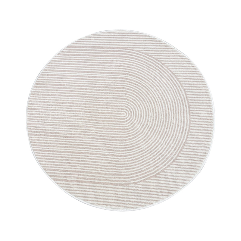 Round beige bathroom rug with a minimalist striped and curved line pattern, placed on a brown floor next to a bathtub, pink towel, and potted plants.