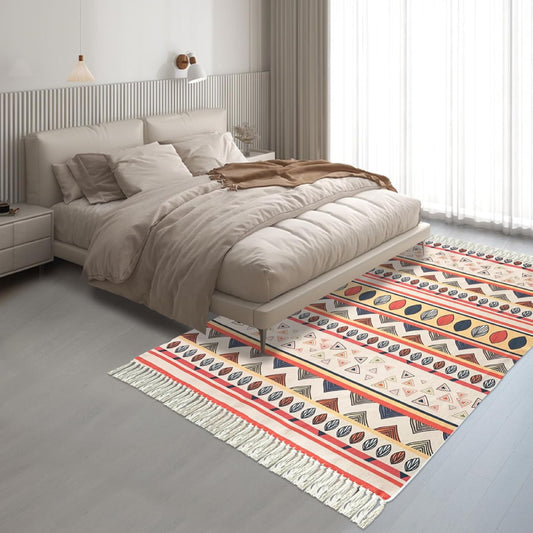 Boho-style handmade rug with colorful geometric and shell patterns, featuring fringe tassels, placed in a bright modern living room with a white sofa, potted plant, and minimalist decor.