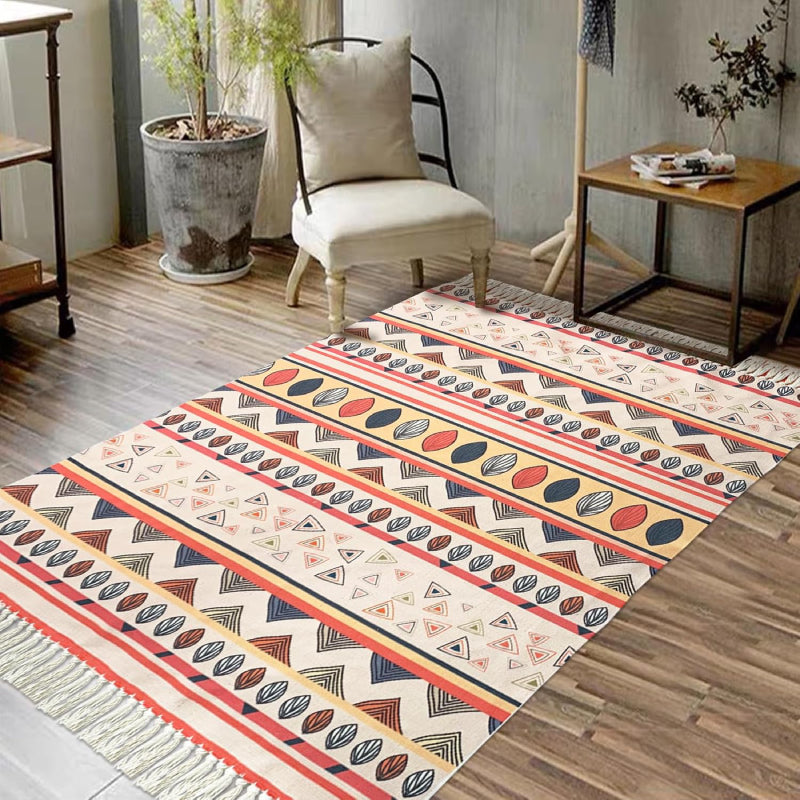 Boho-style handmade rug with colorful geometric and shell patterns, featuring fringe tassels, placed in a bright modern living room with a white sofa, potted plant, and minimalist decor.