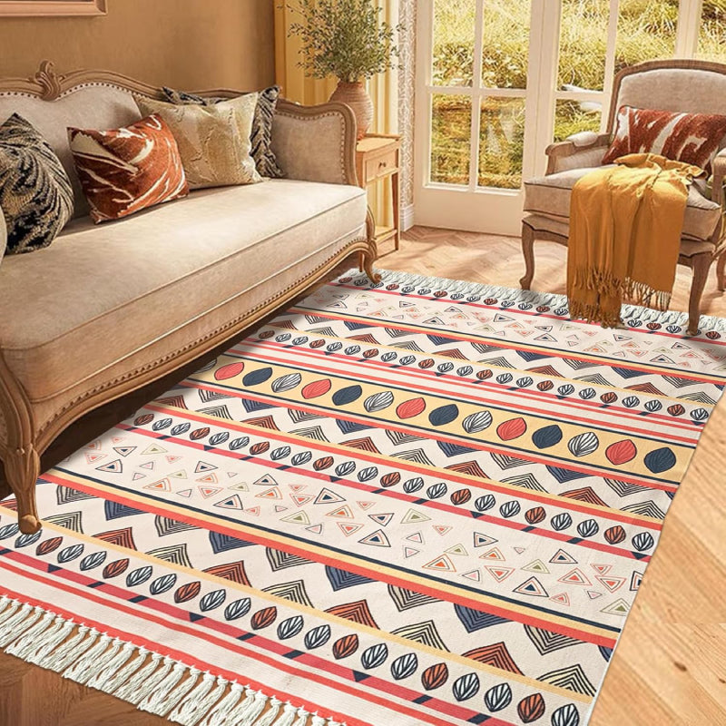 Boho-style handmade rug with colorful geometric and shell patterns, featuring fringe tassels, placed in a bright modern living room with a white sofa, potted plant, and minimalist decor.