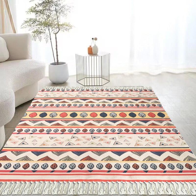 Boho-style handmade rug with colorful geometric and shell patterns, featuring fringe tassels, placed in a bright modern living room with a white sofa, potted plant, and minimalist decor.