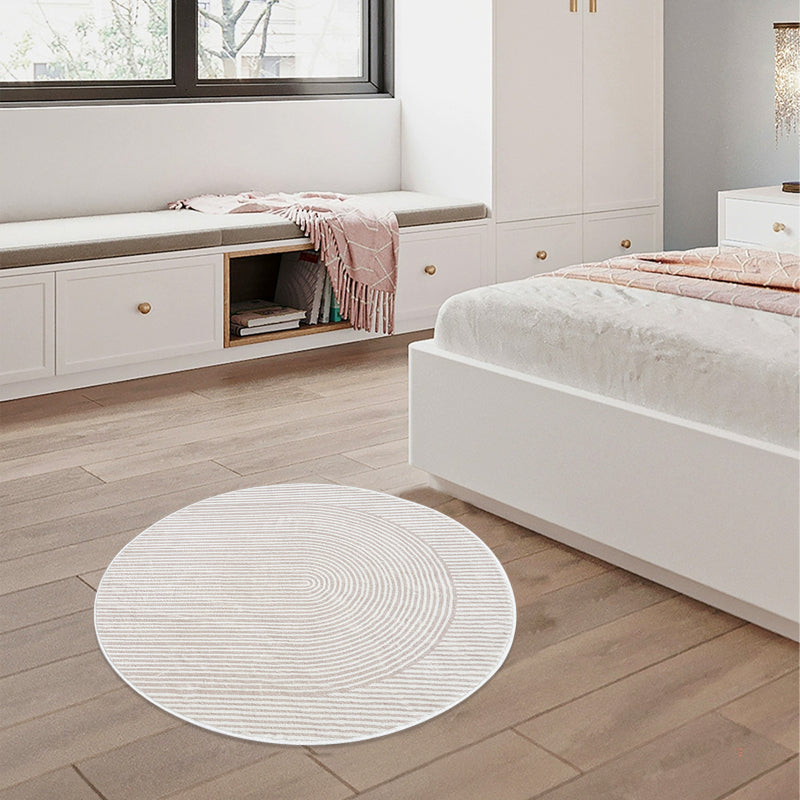 Round beige bathroom rug with a minimalist striped and curved line pattern, placed on a brown floor next to a bathtub, pink towel, and potted plants.