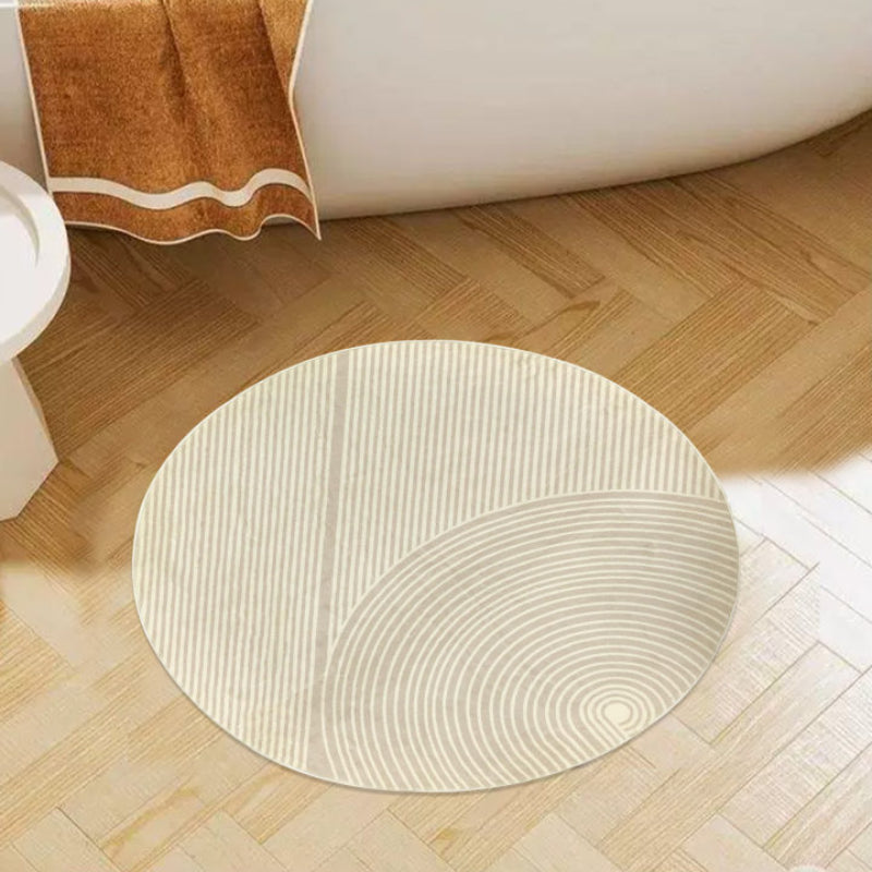 Round beige bathroom rug with a minimalist striped and curved line pattern, placed on a brown floor next to a bathtub, pink towel, and potted plants.