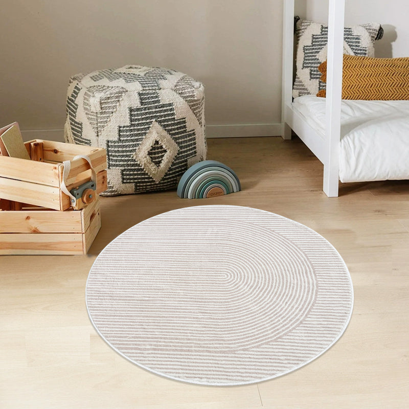 Round beige bathroom rug with a minimalist striped and curved line pattern, placed on a brown floor next to a bathtub, pink towel, and potted plants.